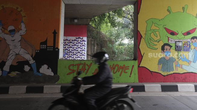 Pengendara sepeda motor melintas di dekat mural bertema COVID-19 di kolong Jalan Tol Lingkar Luar Jakarta, Cipayung, Jakarta Timur, Jumat (3/9/2021). [Cariberita.co.id/Angga Budhiyanto]